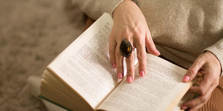 Ein offenes Buch im Schoß einer Frau, die Seiten umzublättern scheint. An ihren Händen trägt sie rosafarben-lackierte Fingernägel und einen großen Ring.